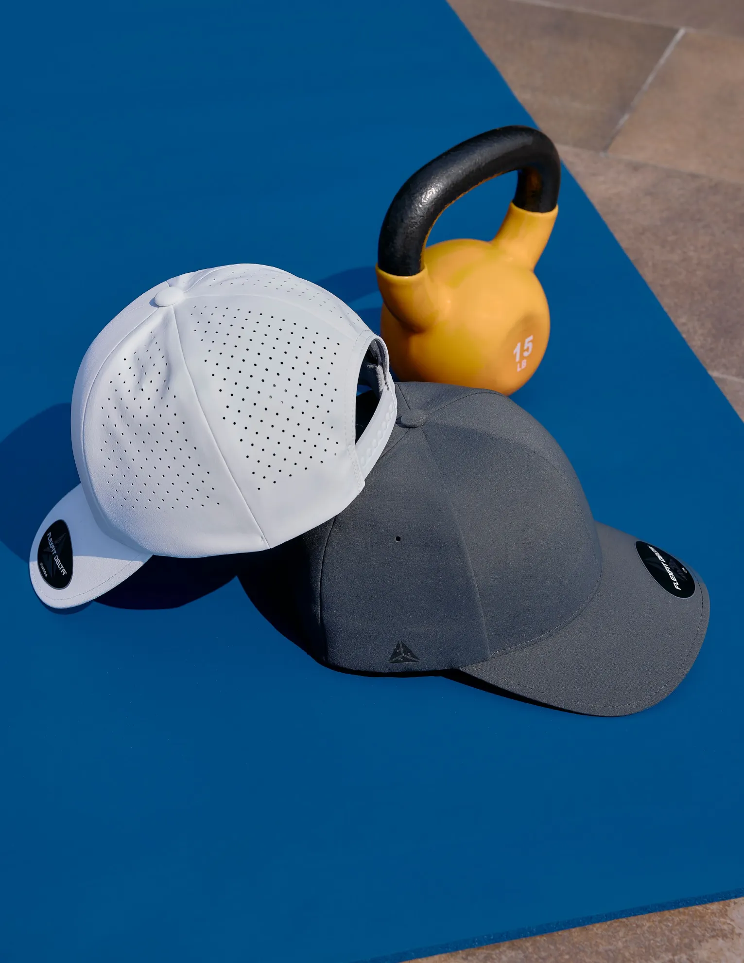 Gorras Flexfit Delta blanca perforada y gris oscuro junto a una pesa rusa sobre una colchoneta azul