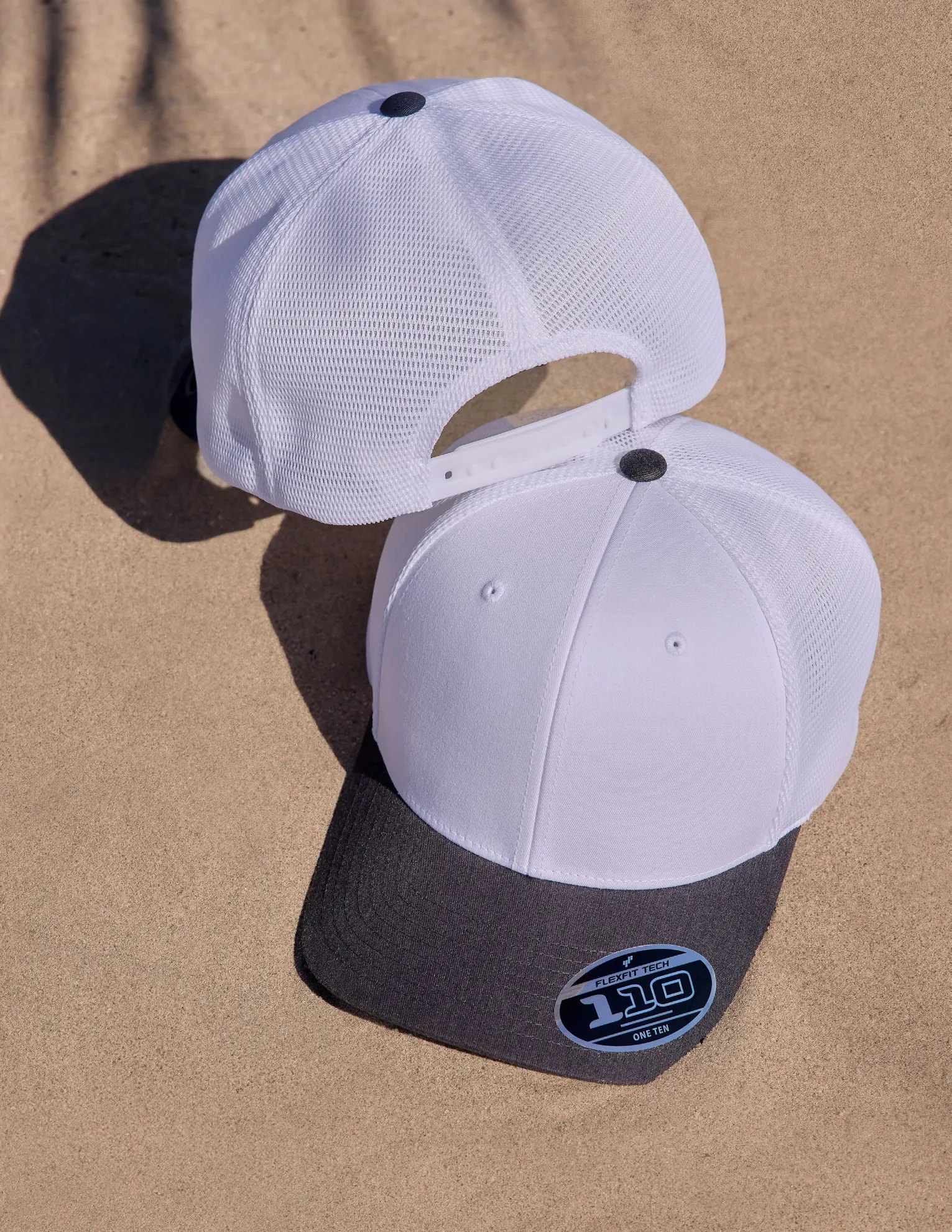 Dos gorras Flexfit 110 blancas con malla y visera gris sobre la arena