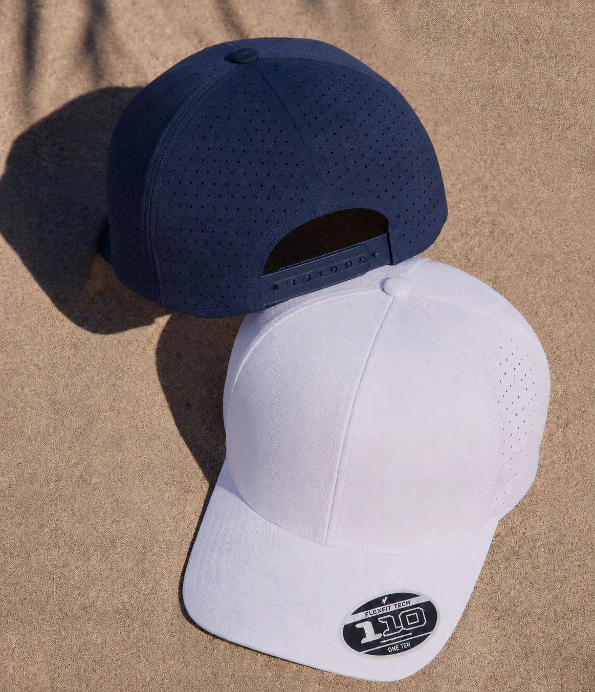 Dos gorras Flexfit 110 perforadas, una azul marino y otra blanca, sobre la arena